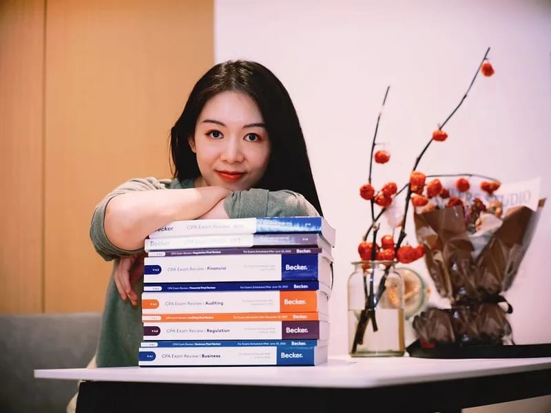 杨同学手臂搭在 8 本 Becker USCPA 教材栈上的全科通关合影